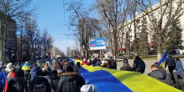 Live-Aufnahmen zeigen Menschen, die ein Banner in den Farben der ukrainischen Flagge halten, während sie am 13. März 2022 in Cherson, Ukraine, gegen die russische Invasion in der Ukraine protestieren, in diesem Standbild aus einem von Reuters erhaltenen Social-Media-Video.
