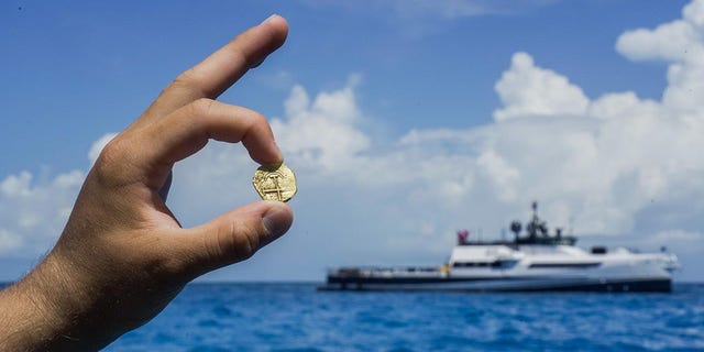 Ein Entdecker hält eine Goldmünze, die auf den Bahamas gefunden wurde, wo das Erkundungsboot der Allen von weitem zu sehen ist.